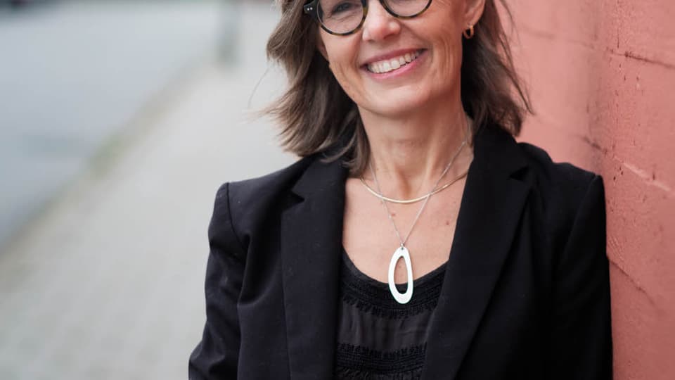 En svartklädd Susanna Hedenborg med glasögon står mot en husfasad och ser glad ut